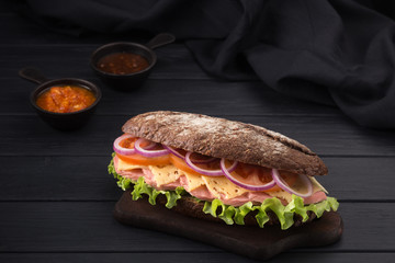 Delicious sandwich on a black wooden table. Sandwich with meat, ham, cheese, lettuce, tomatoes, onion, sauce. Close-up shot. Sandwich bread tomato, lettuce and yellow cheese. Free space for text.
