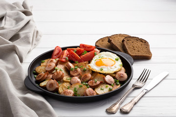 Close-up shot of fried potatoes with sausages and egg. Breakfast. Potatoes with sausages, cut into slices and fried in oil, in rustic style. Frying pan with tasty cooked egg and sausages on table