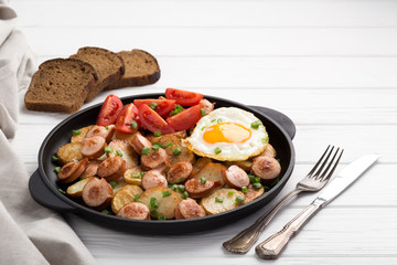 Close-up shot of fried potatoes with sausages and egg. Breakfast. Potatoes with sausages, cut into slices and fried in oil, in rustic style. Frying pan with tasty cooked egg and sausages on table
