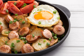 Close-up shot of fried potatoes with sausages and egg. Breakfast. Potatoes with sausages, cut into slices and fried in oil, in rustic style. Frying pan with tasty cooked egg and sausages on table