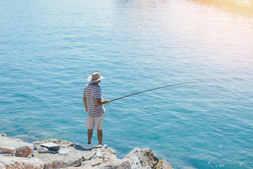Fototapeta na wymiar Fisherman in big hat is fishing on sea rocks alone in sunny day.