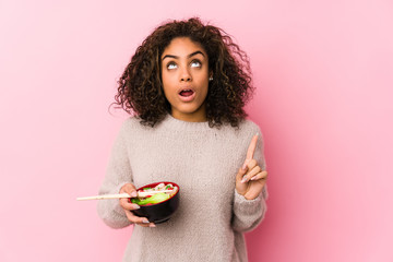 Young african american woman eating noodles pointing upside with opened mouth.