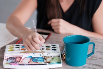 Fototapeta premium Unrecognizable woman mixing watercolors with her thin brush. There's a cup of water next to her paint that she can use to clean her brush.