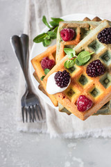A stack of homemade Belgian waffles with frozen berries, mint and natural greek yogurt on a white plate, light grey stone background. Top view.