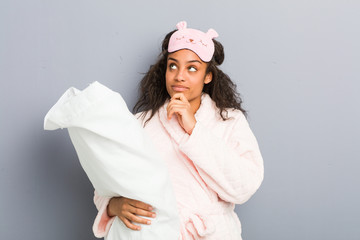 Young african american woman wearing a pajamas and a sleep mask holding a pillow looking sideways with doubtful and skeptical expression.