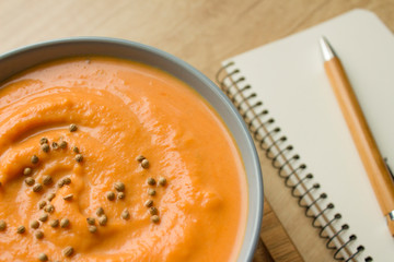 Top view of pumpkin soup in blue bowl on wooden background with recipe book and wooden pen with copy space