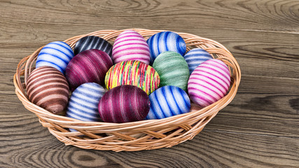 Colored striped ornate Easter eggs in wicker basket on wood. Empty eggshells hand-decorated by glued colorful cotton sewing yarn. Traditional egg shells decoration symbolize Jesus Christ resurrection.