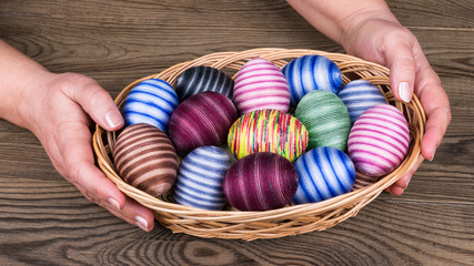 Female hands holding Easter eggs in wicker basket on wooden table detail. Ornate empty eggshells wrapped in colorful cotton thread. Traditional holiday egg shells decorated by glued thin sewing yarn.