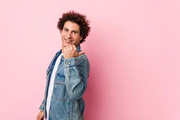 Curly mature man wearing a denim jacket against pink background points with thumb finger away, laughing and carefree.