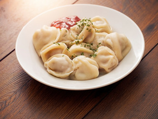 Portion of Russian classic big size dumplings on a white plate with tomato ketchup and herbs. Dark wood table surface.