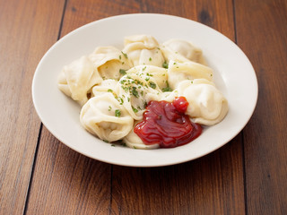 Portion of Russian classic big size dumplings on a white plate with tomato ketchup and herbs. Dark wood table surface.