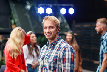 Handsome caucasian blond man looking at camera on the background of dancing people in cozy garden at evening.