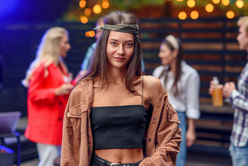 Beautiful stylish caucasian brunette woman in a bandanna looks at the camera while her friends dancing in colored disco light during evening party. Outdoors summer party with dj.