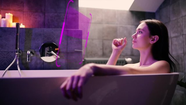 Cute girl takes a bath and using augmented reality futuristic hologram to control the audio player. Young beautiful woman lying in the bathtub and enjoying listening to music