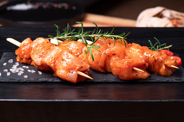 Raw chicken skewers in marinade with spices on a black plate and on a wooden table. Raw marinated...