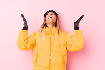 Young caucasian woman wearing a ski clothes in a pink background screaming to the sky, looking up, frustrated.