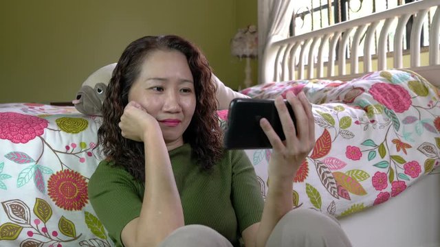 Asian Chinese woman watching video or movie on her smartphone in her bedroom