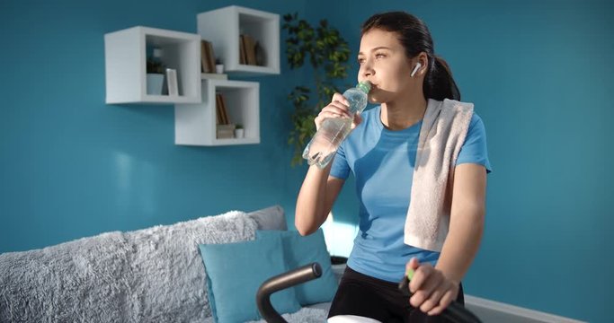 Beautiful slim woman with tied hair knot in black leggings and blue t-shirt drinking water during training on exercise bike. Tired young lady relaxing at home while doing regular workout.