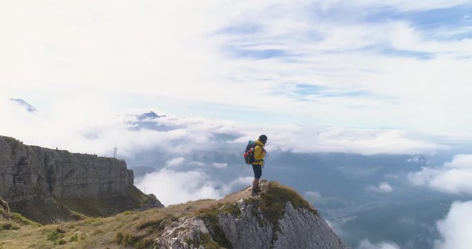 Aerial view. Young man hiking at Seceda mountain raising arms to the sky on the peak at sunrise. Backpack, yellow jacket, boots, beanie. Traveling to puez Odle, Dolomites, Trentino, Italy.