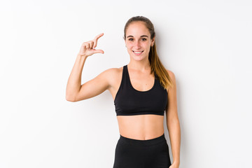 Fototapeta na wymiar Young caucasian fitness woman posing in a white background holding something little with forefingers, smiling and confident.