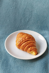 Classic french croissant in a white plate on a blue background. Vertical lifestyle food shot. Close up. Handmade food. Bakery business. Copy space. Natural light.