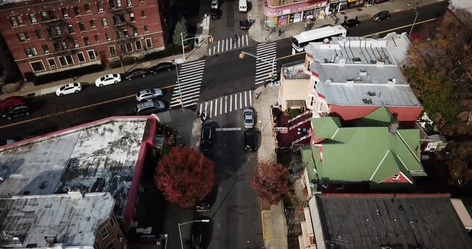 Cars and buses drive on the streets as pedestrians walk on the crosswalk casting shadows on the sidewalk past buildings of Brooklyn with the New York City skyscrapers on the horizon. Aerial Drone