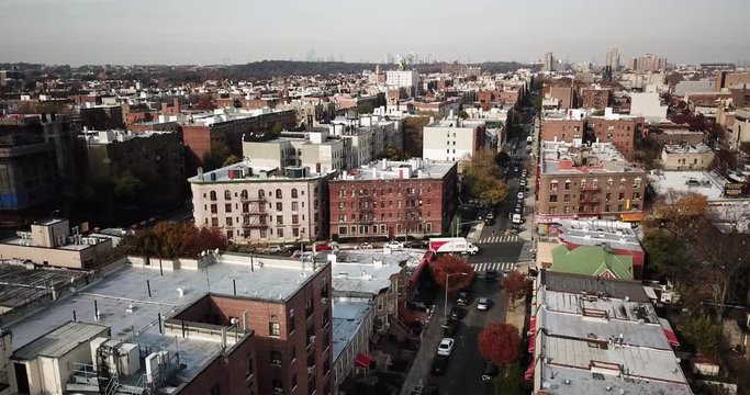 Cars drive on the streets and pedestrians walk on the crosswalk past buildings of Brooklyn with the New York City skyscrapers on the horizon on a cloudy day. Aerial Drone