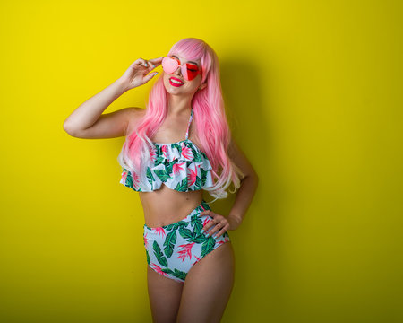 Beautiful girl in a pink wig and colored bikini posing on a yellow background. Woman with artificial long hair and pink glasses in the form of hearts.