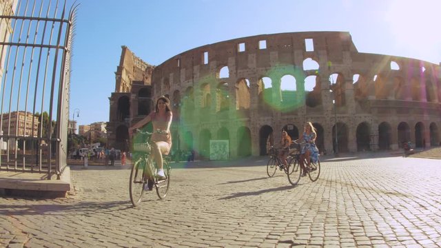 Three happy young women friends tourists riding bikes at Colosseum in Rome, Italy at sunrise.