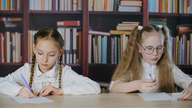 Focused schoolgirls writing exam and cheating cheat sheet in classroom. Bookshelf background. Adorable teenage girls sitting at desk, talking and cheating during exam in school