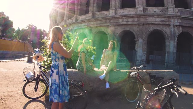 Three happy young women friends tourists with bikes taking videos with smarphone at Colosseum in Rome, Italy at sunrise.