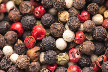 Close-up of assorted peppercorns in black, red, white, and green. Some are intact, while others are cracked, revealing seeds. The mix displays vibrant and earthy tones with a rough, natural texture.