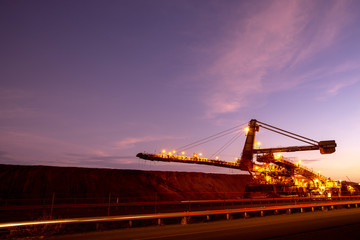 Obraz premium Large mining industry equipment reclaimer machine, conveyor belts moving raw copper, silver, gold, from train load out stockyard to ship load out areas ready to export with sunset at the background 
