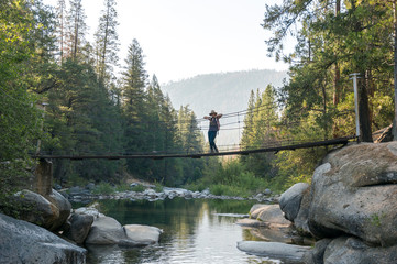 Young woman female hiker in hat stands on suspension bridge over beautiful stream lined with rocks in front of forest and mountains