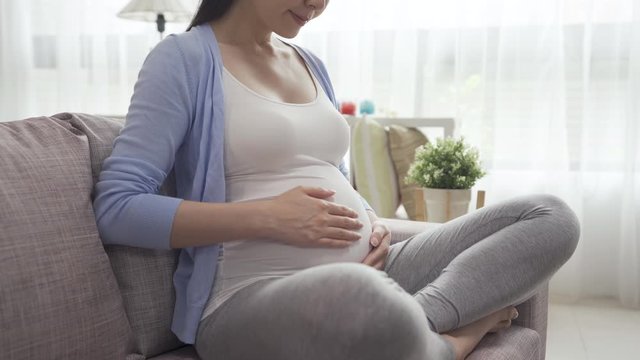 pregnancy and motherhood concept. focus on huge belly of pregnant woman sitting on couch and hands touching in sunlight living room. young future female parent feeling unborn baby in big tummy.