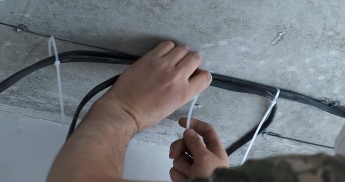 Male electrician stretches high-voltage cable across ceiling of apartment, remodels electrical wiring, twists wires with plastic clamps, works quickly, fastens wires to metal wire, construction