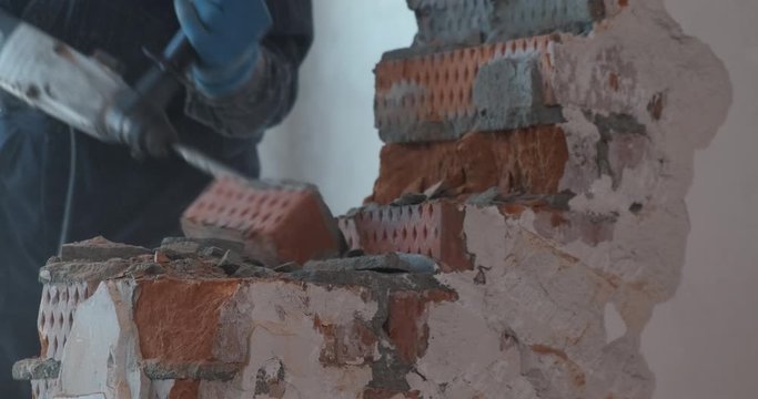 Male builder work with jackhammer, demolishes wall in apartment, pick out bricks from masonry, breaks piece, throws it to side, cleans dust and debris, stones fly in different directions, construction