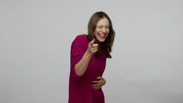 You are ridiculous! Overjoyed amused brunette woman in pullover bursting into laughing and pointing to camera, crying with hilarious laughter, making fun of friend. indoor studio shot gray background