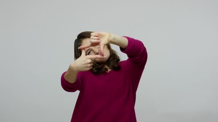 Cheerful brunette woman in pullover looking through photo frame made of fingers, viewing at camera with interest, having fun, pretending to take picture. indoor studio shot isolated on gray background
