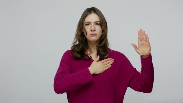 Responsible brunette woman in pullover looking into camera with devoted eyes, making promise honestly with hand up, swearing fidelity, taking oath. indoor studio shot isolated on gray background