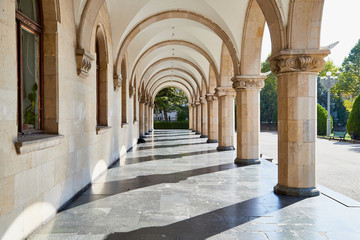 Fototapeta na wymiar Arcade and a corridor of white columns on one side and walls on the other. A passage of marble columns outside and green plants in Park. Background with perspective going into distance