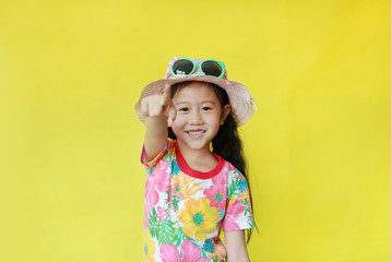 Happy asian girl pointing at camera isolated over yellow background. Kid in floral pattern summer dress and hat with sunglasses. Holiday and summer fashion concept.