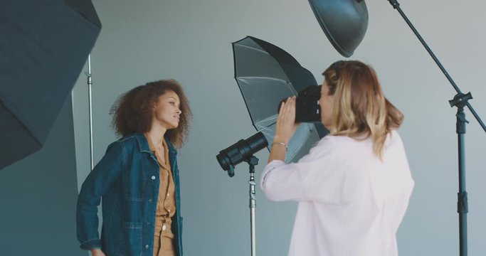 Young professional female photographer taking photos of a beautiful african american woman modeling for a luxury clothing brand, behind the scenes of a studio fashion photoshoot