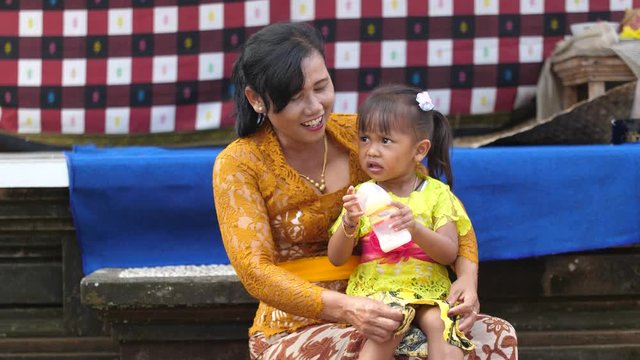 Asian woman in saffron colored lace top with toddler on her lap smiles for the camera. Indonesian lady with her daughter sitting in temple courtyard after religious event and celebration.