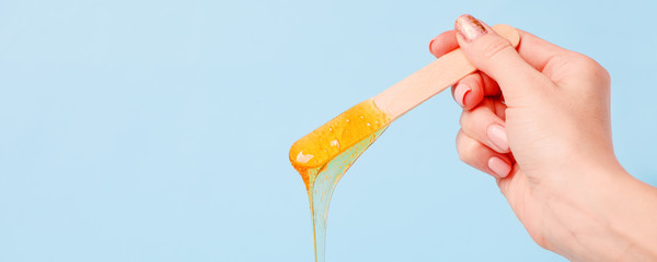Hand holding beauty wax or sugark paste on wooden spatula flowing down into container on blue...