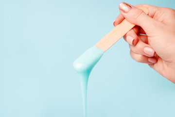 Hand holding beauty wax or sugark paste on wooden spatula flowing down into container on blue background. Advertising beauty industry concept, luxury skincare bodycare depilation idea - obrazy, fototapety, plakaty