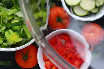 A glass cover from a saucepan is hold in front of washed carved vegetables, leek, couchette, tomatoes and paprika.