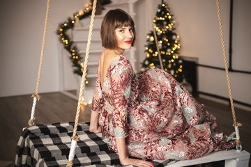 Indoor portrait of confident girl in a long dress posing near christmas tree on the swing.