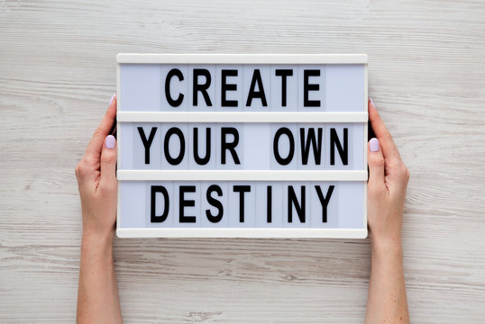 Female hands hold a lightbox with 'Create your own destiny' words over white wooden background, top view. Overhead, from above, flat lay.