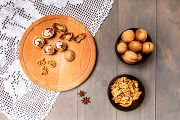 Still life. On a gray table lies a white handmade lace napkin. Round wooden board. In cups whole and peeled walnuts. View from above.